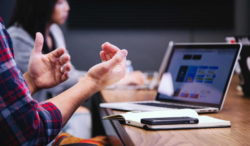 Product Owner in a meeting with hands up in front of apple laptop and iPhone 8 mobile devices